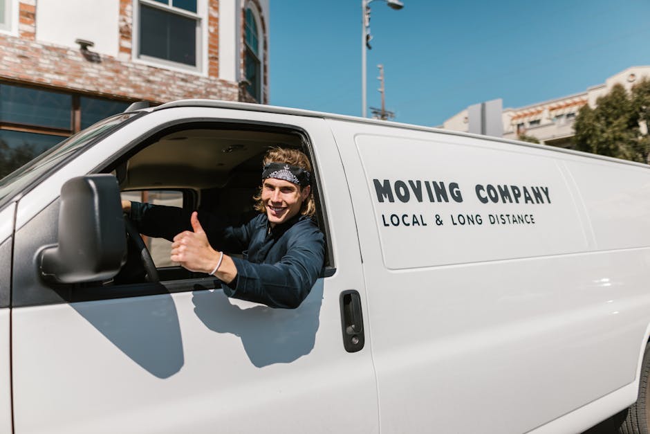 A male mover with long, wavy hair and a grey bandana, sitting in the driver's seat of a white van branded with 'Moving Company Local & Long Distance,' inside a housing environment. The important part 