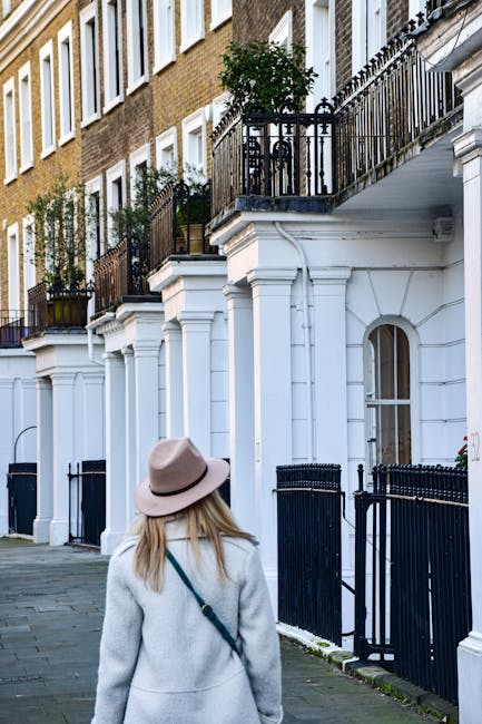 A woman wearing a beige wide-brimmed hat and a light grey coat is walking along a street lined with white terraced houses featuring black wrought-iron balconies and railings. She is carrying a dark shoulder bag, and her blonde hair is visible beneath the hat. The pavement consists of grey stone slabs, and there are potted plants on some of the balconies. The scene is well-lit, indicating daylight, and represents a residential area suitable for house removals and home relocation services, with the buildings' features and street layout visible as part of the moving process context offered by Man with Van Soho.