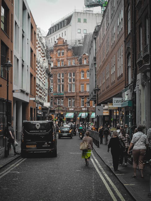 A narrow city street in Soho during daylight, with pedestrians walking on both sides of the road, some carrying bags and luggage. The street is surrounded by tall multi-storey buildings featuring a mix of modern and historic architecture with brick facades and large windows. Parked cars, including a black van with the branding 'download the app,' are visible along the curb, while a few vehicles are driving through the street. The pavement is lined with small shops, cafes, and offices, with visible signage, some of which relate to home services. The lighting is natural, illuminating the urban environment, and the scene captures typical busy street activity associated with city house relocations and furniture transport logistics, with [COMPANY_NAME] possibly involved in the background activities of removals and moving services in Soho.
